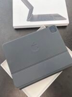 White box showing a folded laptop accessory; in front, a dark gray Apple device with a logo in a protective case.