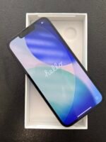 Smartphone in an open white box on a dark surface, screen on with blue abstract wallpaper and a notch at the top edge