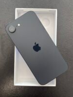 Black smartphone (iPhone) with dual‑camera system resting in its white box, viewed from above showing the back and Apple logo in dark gray.