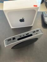 White Apple Mac mini box with black Apple logo on the front, resting on a desk beside a silver docking hub and payment terminal