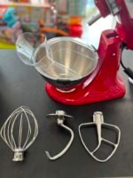 Red stand mixer with a stainless steel bowl and three attachments (whisk, beaters, dough hook) on a dark countertop; blurred colorful store shelves in the background.