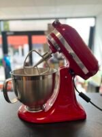 Red stand mixer with a stainless steel mixing bowl on a dark countertop, in a bright shop background