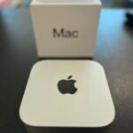 White Mac product box on a dark table with a foreground card featuring the Apple logo (slightly blurred).