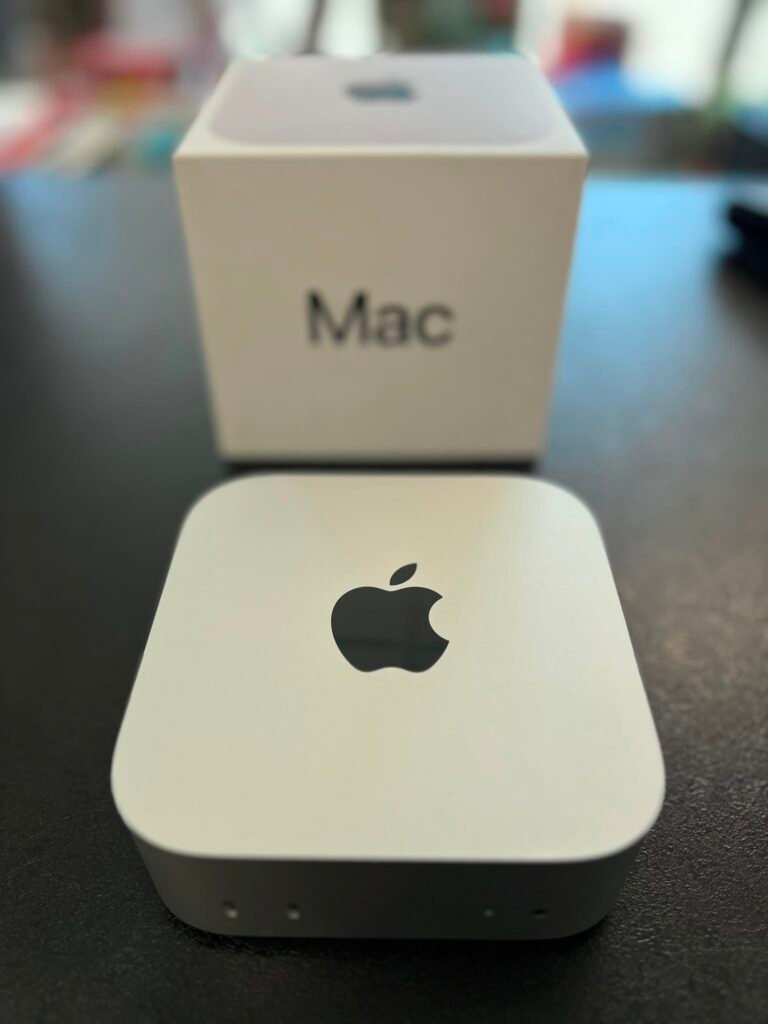 White Mac product box on a dark table with a foreground card featuring the Apple logo (slightly blurred).
