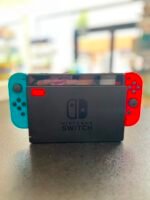 Close-up of a Nintendo Switch with blue left Joy-Con and red right Joy-Con on a table, screen off, blurred store background.