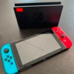Nintendo Switch with blue left and red right Joy-Cons resting on a table, screen off, background blurred.