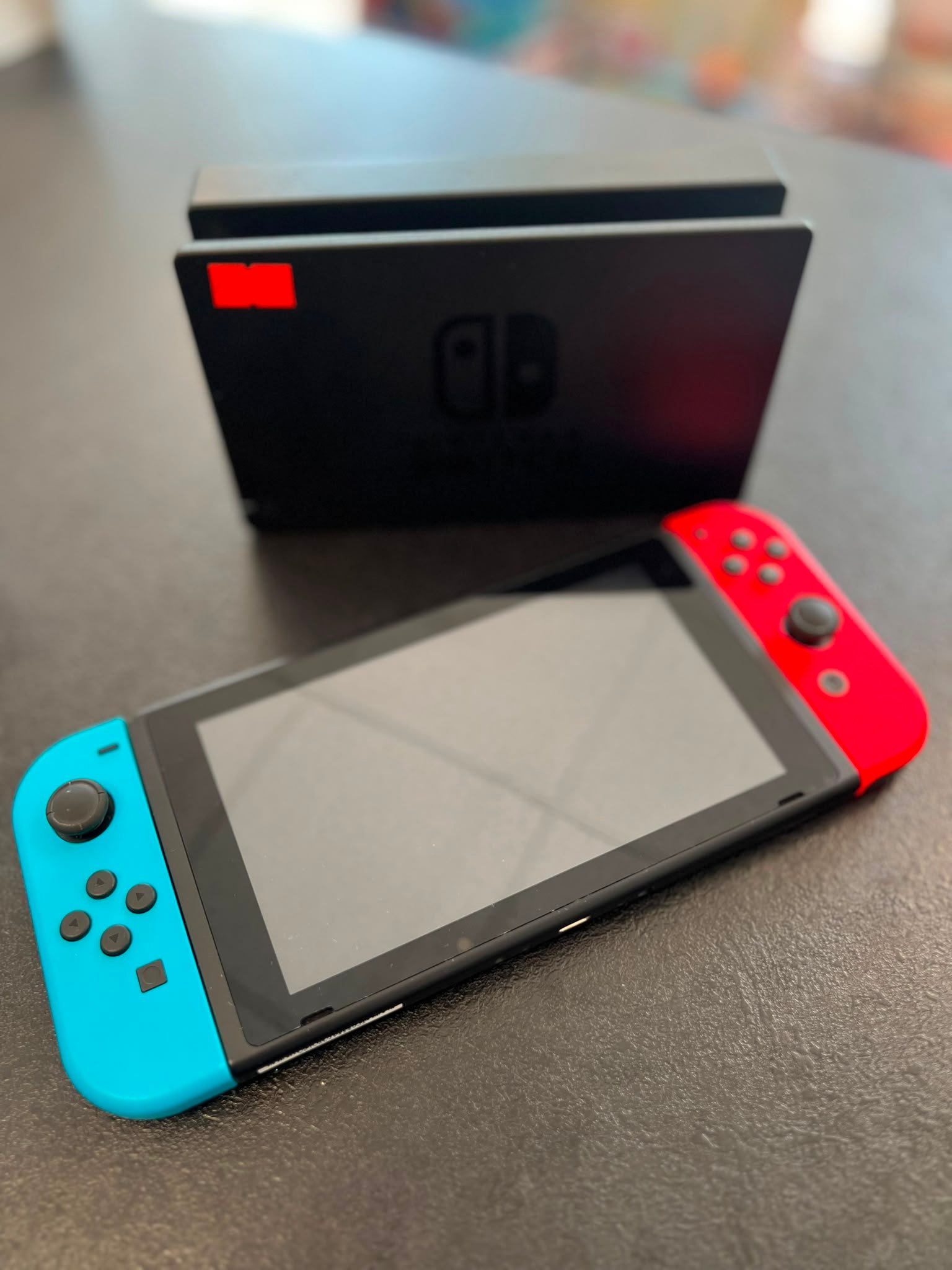 WhatsApp Image 2026-07-14 at 124414 - Radau Viska Nintendo Switch with blue left and red right Joy-Cons resting on a table, screen off, background blurred.