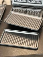 Close-up of metallic ridged grill plates from several sandwich makers stacked on a dark countertop