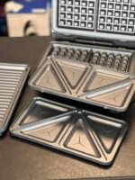 Stacked metal lunch tins with triangular compartments on a dark countertop, with a blurred grid-pattern appliance in the background.