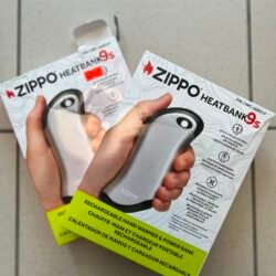Hands holding two Zippo HeatBank 9s rechargeable hand warmer boxes, gray device visible, bright yellow warning labels on the boxes.