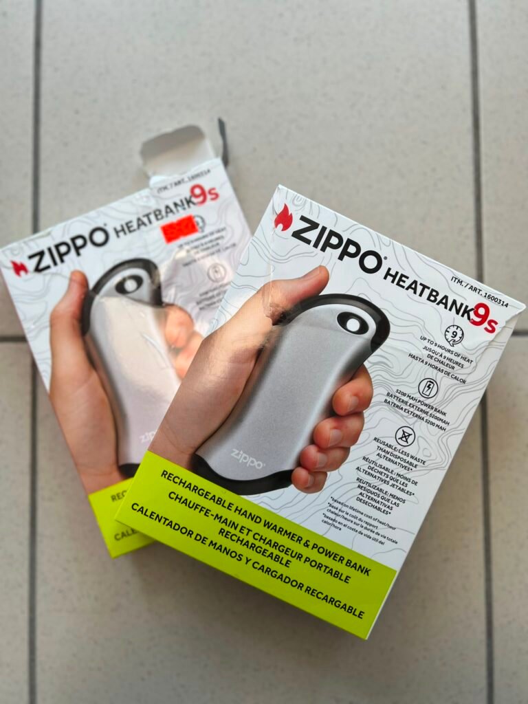 Hands holding two Zippo HeatBank 9s rechargeable hand warmer boxes, gray device visible, bright yellow warning labels on the boxes.