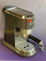 Close-up of a chrome DeLonghi espresso machine top with a ridged warming surface and attached portafilter edge streaked by reflections.