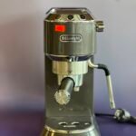 Front view of a DeLonghi espresso machine head with chrome controls and a red sticker, highlighting the machine's top panel and portafilter area.