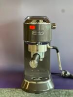 Front view of a DeLonghi espresso machine head with chrome controls and a red sticker, highlighting the machine's top panel and portafilter area.