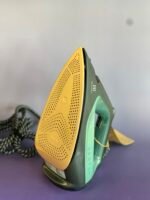Close-up of an iron with a yellow perforated soleplate and green body, resting on a stand.