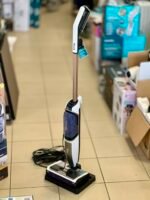 Shark steam mop standing upright on a display base in a store, with a coiled cord on the tiled floor behind it.]