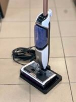 Vertical garment steamer with white and brown body, purple water reservoir, and upright wand attached to its base on a tiled floor. The power cord is coiled in the background.