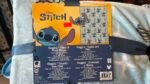 Disney Stitch plush toy packaging with orange background and a silicone Stitch graphic; price tag shows 25.00 and a patterned insert with Stitch images on gray panel