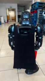 Back view of a humanoid robot with dark metallic armor and a black fabric covering its torso in a workshop