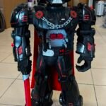 Life-size Darth Vader-style robot figure in a workshop, holding a red lightsaber and featuring red accents and a metallic chest piece.