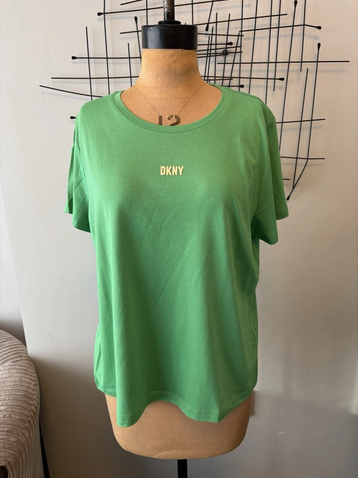 s-l1600 5 - Radau Viska Green DKNY T-shirt displayed on a fashion mannequin with an abstract black metal wall sculpture behind it.