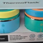 Two-tone ThermoFlask insulated food jars in a display box with teal and blue bases and yellow/orange lids, brand name visible on top box edge.