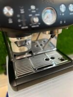 Close-up of an espresso machine front: portafilter attached, gauge visible, and metal drip tray in view.