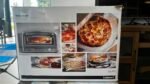 Box showing a 'Pizza Oven' by Cuisinart with collage of pizzas and an oven image on the front panel, product packaging visible in a showroom.