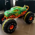 Colorful green toy monster truck with oversized black tires and bright orange suspension on a tiled floor indoors, resembling a display model.