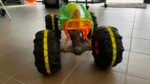 Green toy RC monster truck with oversized black tires and orange bumper on a tiled showroom floor, indoors.
