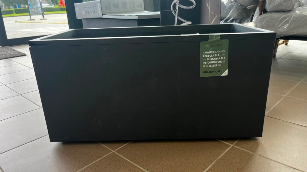 Large black outdoor storage box with a green Deroma Millennium tag attached to the side of the box in a showroom setting