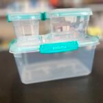 Transparent two-compartment pill organizer with turquoise lids connected side by side on a blue base; blurred background behind it.