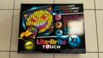 Lite-Brite toy box open to reveal colorful pegs and a small handheld kit on a black glossy box with 'Lite-Brite' logo and 'TOUCH' text nearby.