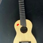 Full view of a light-colored guitalele lying on a black table, showing the soundhole with a label and a red sticker near the lower bout.