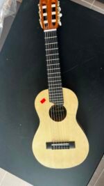 Full view of a light-colored guitalele lying on a black table, showing the soundhole with a label and a red sticker near the lower bout.