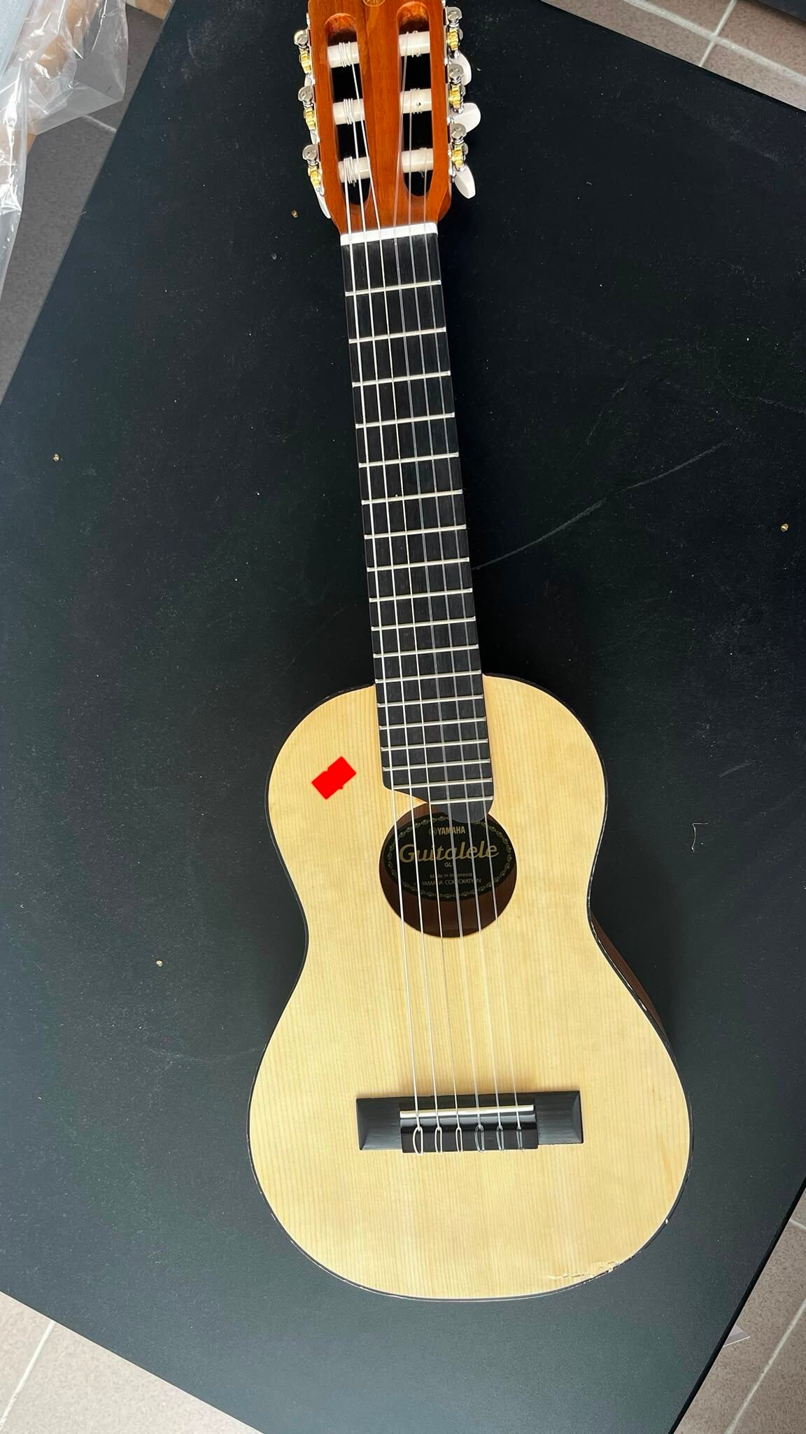 WhatsApp Image 2026-08-05 at 103204 - Radau Viska Full view of a light-colored guitalele lying on a black table, showing the soundhole with a label and a red sticker near the lower bout.