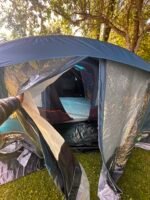 Open blue-gray camping tent set up in a wooded campsite, with trees and grass around and tent flaps partially opened.
