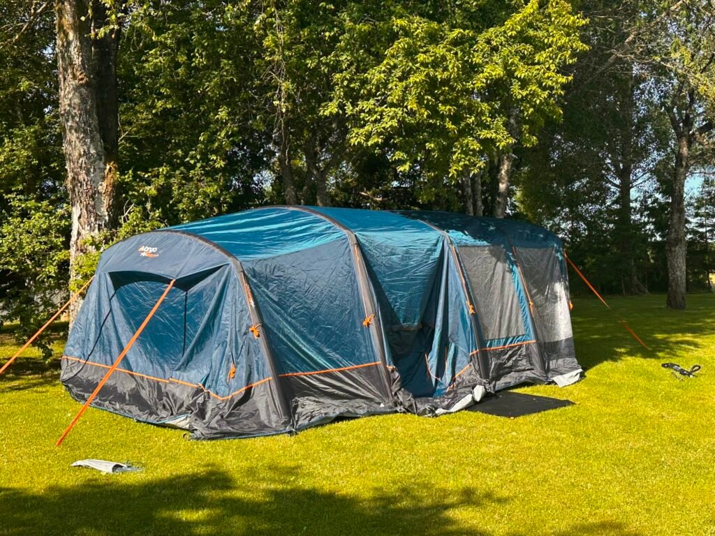 Blue and gray Camping tent pitched on a sunny grassy lawn with tall trees behind it.