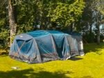Blue and gray Camping tent pitched on a sunny grassy lawn with tall trees behind it.