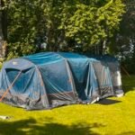Blue and gray Camping tent pitched on a sunny grassy lawn with tall trees behind it.