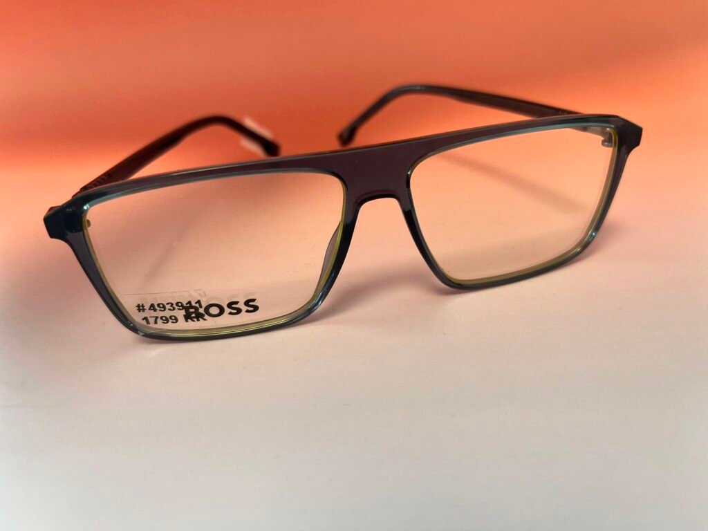 Close-up of a black plastic eyeglass frame with clear lenses and a price sticker on the left lens against an orange background, brand tag reading BOSS nearby.