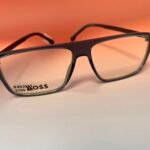 Close-up of a black plastic eyeglass frame with clear lenses and a price sticker on the left lens against an orange background, brand tag reading BOSS nearby.