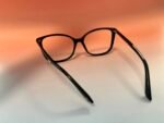 Close-up of black eyeglasses with rounded rectangular frames resting on a light surface against an orange gradient background.