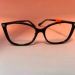 Black plastic eyeglasses with a small orange tag on the right temple, against a peach background.