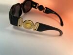 Close-up of black sunglasses with a gold circular emblem on the temple, resting on a light surface.