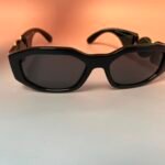 Close-up of black oversized sunglasses with gold accents on a peach to white gradient background