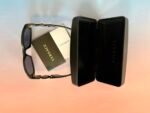 Black sunglasses with gold hinges placed next to a Versace tag and a black Versace case on a peach-to-pink gradient background.
