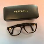 Black Versace glasses case with a gold logo, beside brown tortoiseshell eyeglasses on a light background.