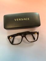 Black Versace glasses case with a gold logo, beside brown tortoiseshell eyeglasses on a light background.