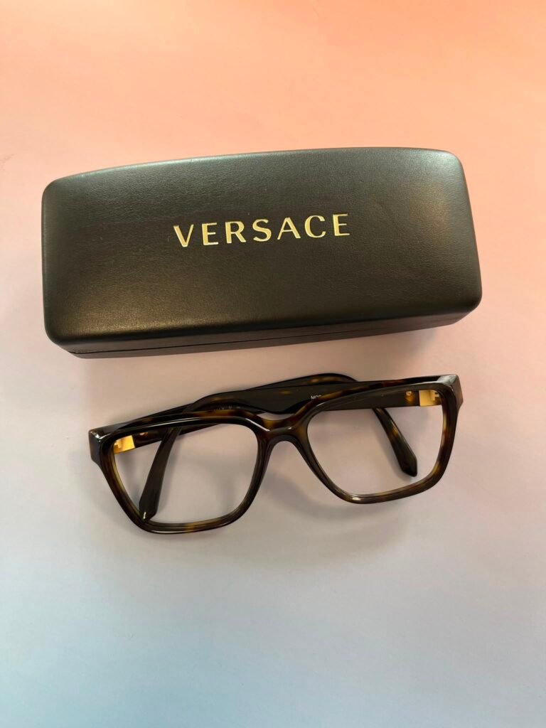 Black Versace glasses case with a gold logo, beside brown tortoiseshell eyeglasses on a light background.